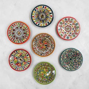 May include: A collection of six circular ceramic plates featuring intricate hand-painted geometric and floral patterns in vibrant colors including red, orange, green, blue, and yellow, arranged in a circular formation on a neutral surface.