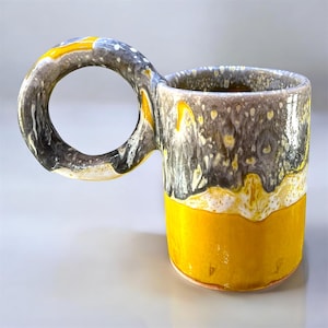 Peut inclure: Une tasse en céramique dotée d'une grande anse circulaire. Le corps de la tasse présente un émail bicolore avec une base jaune vif unie qui se transforme en une section supérieure grise et blanche mouchetée et texturée, créant un effet goutte.