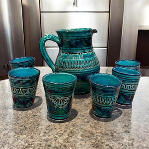 Peut inclure: Un pichet en céramique et cinq gobelets assortis arborant un émail bleu sarcelle et bleu foncé. Chaque pièce est ornée de motifs géométriques peints à la main, incluant des triangles répétitifs et des motifs floraux sur l'extérieur.