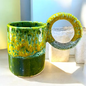 Peut inclure: Une tasse en céramique avec un corps cylindrique, une base vert foncé brillant et une partie supérieure tachetée de jaune et de vert. La tasse possède une grande anse circulaire en forme d'anneau avec une finition émaillée jaune et blanche.