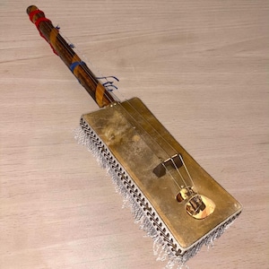 Peut inclure: Une guitare à trois cordes fabriquée à partir d'une boîte à cigares avec un long manche en bois entouré de bandes de tissu rouge et bleu. Le corps rectangulaire présente une finition dorée, un chevalet sombre et une frange argentée.