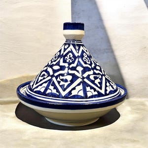 Peut inclure: Un tajine en céramique composé d'un couvercle conique et d'une base large. L'extérieur est orné d'un motif géométrique bleu et blanc peint à la main, composé de losanges entrelacés et de motifs floraux, surmonté d'une petite poignée cylindrique.