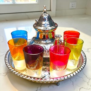 May include: A silver-toned metal teapot with a lid sits on a matching round silver tray, surrounded by six small glass cups in vibrant colours including blue, orange, purple, red, yellow, and green, each featuring gold filigree patterns.