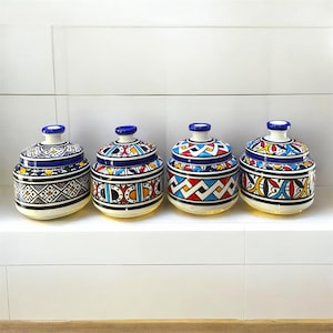 Peut inclure: Quatre pots en céramique avec couvercles, chacun présentant un motif géométrique peint à la main dans des tons vifs de bleu, rouge, jaune et noir. Les pots ont une forme arrondie avec un petit bouton sur chaque couvercle et une base blanche.