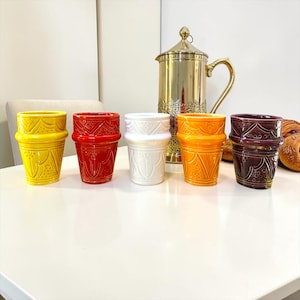 Peut inclure: Un ensemble de cinq tasses en céramique jaune, rouge, blanche, orange et violet foncé, chacune ornée d'un motif géométrique en relief. Derrière les tasses se trouve une grande carafe en métal doré brillant avec une anse et un couvercle.