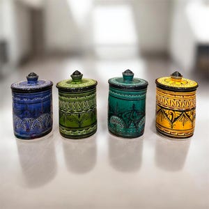 May include: Four cylindrical ceramic canisters with lids, each featuring a distinct color: blue, green, teal, and yellow. Every canister is decorated with intricate black hand-painted patterns, including geometric bands and floral-inspired motifs.