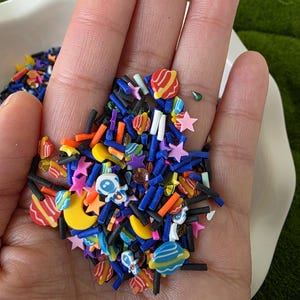 Könnte beinhalten: Eine Handvoll bunter Polymer Clay Streusel in verschiedenen Formen, darunter Sterne, Planeten, Monde und rechteckige Stäbchen. Die Streusel sind in den Farbtönen Blau, Orange, Gelb, Rosa und Schwarz gehalten. Die Streusel befinden sich in einer weißen Schüssel.