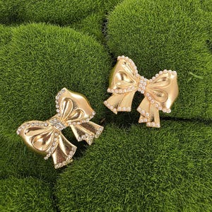 May include: Two gold-colored bow-shaped hair clips with rhinestone and pearl accents. The clips are displayed on a bed of green moss. The bows have a shiny, metallic finish and are intricately detailed.