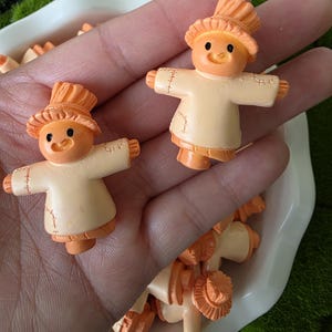May include: Two small, orange and cream-coloured scarecrow figurines. Each figurine has a hat, stitched details, and outstretched arms. The figurines are in a white dish, with a green moss background.