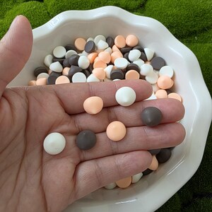 May include: A white, ruffled-edge bowl filled with small, round candies in white, brown, and peach colors. Some candies are held in a hand, showcasing the variety of colors and textures. The background is a bed of green moss.