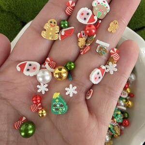 Puede incluir: Una colección de pequeños adornos navideños para manualidades que incluye miniaturas de hombres de jengibre, caras de Papá Noel, árboles de Navidad verdes, copos de nieve blancos, bastones de caramelo y cuentas redondas variadas.