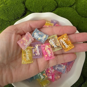 May include: A collection of colorful, miniature candy-shaped resin charms. Each charm is a different color, including pink, blue, yellow, and green, with the word "Sweet" printed on each. The charms are held in a white dish.