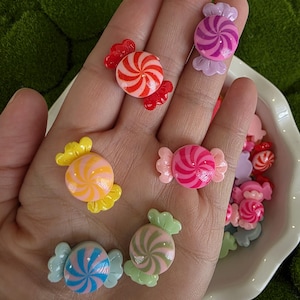 May include: A collection of colorful, miniature candy-shaped embellishments. The candies feature a swirl design in various colors, including red, pink, blue, green, and yellow. Each candy has a decorative wrapper.