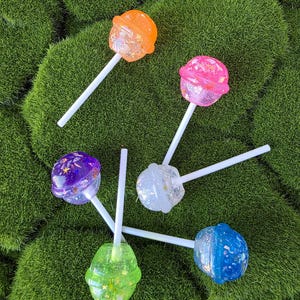 Puede incluir: Una colección de piruletas coloridas con purpurina e inclusiones en forma de estrella. Las piruletas son de color naranja, rosa, morado, blanco, verde y azul, cada una con un palito blanco. El fondo es una superficie verde texturizada.