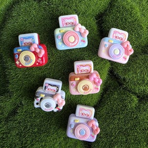May include: Six small camera-shaped charms in red, light blue, pink, brown, white with black spots, and purple. Each charm features a pink bow, a lens with the word Sweet, and a top screen displaying a white cat character wearing red glasses.