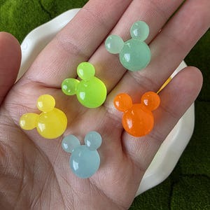 May include: Five colorful, translucent Mickey Mouse-shaped beads in yellow, green, orange, blue, and light green. The beads are smooth and rounded, with a glossy finish. The beads are small and appear to be made of resin or a similar material.