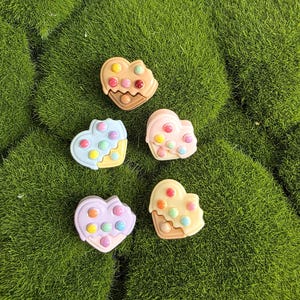 May include: Five heart-shaped charms in pastel colors, each featuring a textured surface with small, raised circular dots in various shades. The charms are arranged in a cluster, displaying light blue, light pink, lavender, and tan color variations.