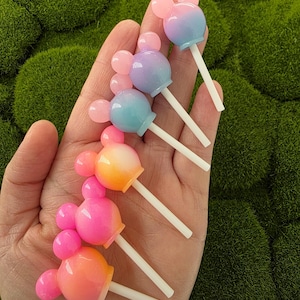 Puede incluir: Una colección de piruletas coloridas con forma de ratón. Cada piruleta presenta un degradado de colores, incluyendo rosa, naranja, azul y morado, con un palito blanco. Las piruletas están dispuestas sobre un fondo de musgo verde.