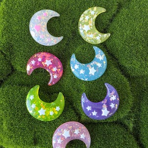 May include: Seven crescent moon-shaped resin charms in various colors, including pink, green, blue, and purple. Each charm is filled with glitter and white star-shaped accents, set against a green moss background.