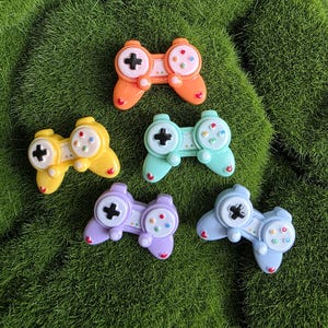 May include: Five small, glossy game controller charms in orange, yellow, mint green, lavender, and light blue. Each controller features a black directional pad on the left, four small colored buttons on the right, and a tiny red heart at the base.