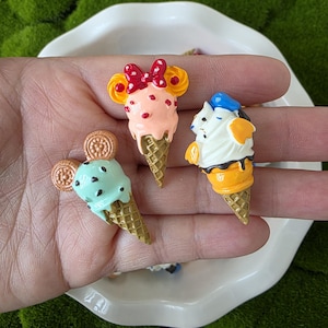 May include: Three miniature ice cream cone figurines. One has pink ice cream with a red bow, another has mint green ice cream with a cookie, and the third has white ice cream with a blue hat. All have waffle cone bases.