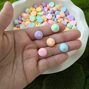May include: A hand holds several pastel-colored, round candies, with more in a white, scalloped bowl. The candies are in shades of pink, yellow, blue, green, and purple. The background is a bed of green moss.