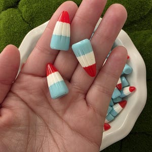Puede incluir: Una colección de piezas de arte de uñas coloridas con forma de mini polos. Cada pieza presenta una punta roja, una franja blanca y una base azul claro. El arte de uñas se muestra en un plato blanco.