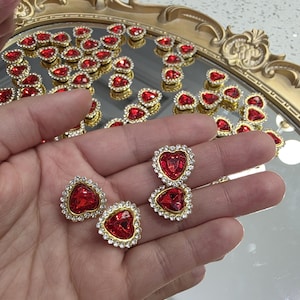 Könnte beinhalten: Rote herzförmige Strass-Verzierungen mit goldfarbenen Metalleinfassungen und klaren Strass-Rändern. Die Verzierungen werden auf einer reflektierenden Oberfläche präsentiert, für Schmuck oder Bastelprojekte.