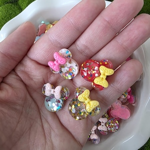 May include: Close-up of several clear resin mouse-shaped charms with glitter and bow accents. The bows are pink, yellow, and light blue. The charms are in a white bowl, with a green background.