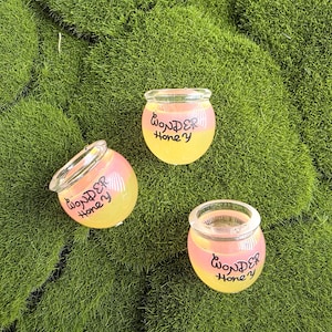 Könnte beinhalten: Drei kleine, runde, durchsichtige Glasgefäße mit einem rosa-gelben Farbverlauf. Jedes Gefäß ist auf der Vorderseite mit dem schwarzen Schriftzug "WONDER Honey" bedruckt. Die Gefäße haben eine weite, offene Randöffnung und einen abgerundeten Boden.