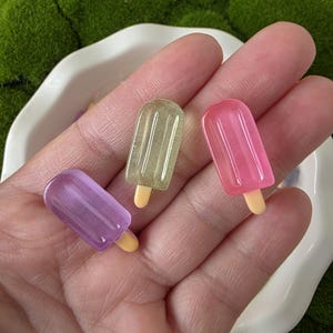 Pode incluir: Três pingentes de picolé em miniatura em tons de roxo, verde e rosa. Cada pingente tem um pequeno palito amarelo. Os picolés são dispostos em um prato branco, com um fundo verde semelhante a musgo.