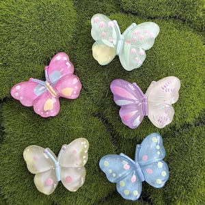 Op de afbeelding: Vijf kleurrijke vlindervormige decoraties. De vlinders zijn in tinten roze, paars, blauw en geel, met verschillende patronen en details. Ze zijn gerangschikt op een bed van groen mos.