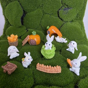 May include: A collection of miniature Easter-themed figurines on a bed of green moss. The figurines include white bunnies, a carrot house, a pumpkin house, a wooden cart, a cabbage, a fence, and pumpkins. The scene evokes a whimsical, springtime feel.