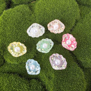 May include: Seven colorful seashell-shaped resin charms with a pearl in the center. The charms are in shades of pink, yellow, green, blue, and purple, with glitter accents. The charms are set against a background of green moss.