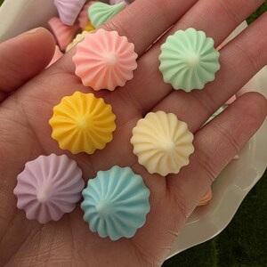 May include: A collection of colorful, swirl-shaped resin embellishments in pastel shades of pink, yellow, blue, green, and lavender. The decorative pieces are arranged in a hand and in a white bowl, with a green moss background.