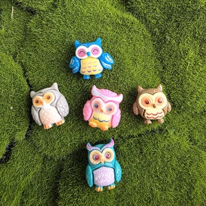 May include: Five small owl figurines in various colours. One is blue with yellow accents, one is grey, one is pink with yellow, one is brown, and one is teal with purple. Each owl has distinct facial features and textured wing details on their bodies.