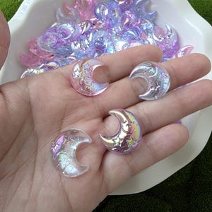 Può includere: Abbellimenti iridescenti a forma di mezzaluna con disegni di stelle e vortici. I pezzi decorativi sono nelle tonalità del rosa, del viola e del trasparente, con un effetto olografico scintillante. Una manciata di ciondoli è tenuta nella mano di una persona.