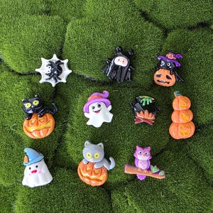 Può includere: Una collezione di figurine in miniatura a tema Halloween. Le figurine includono un gatto nero su una zucca, un fantasma, un ragno, una strega, un gufo e un calderone. Le figurine sono in vari colori, tra cui arancione, nero, bianco e viola.