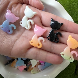 Op de afbeelding: Een verzameling kleine, katvormige versieringen in verschillende kleuren, waaronder wit, zwart, roze en een verloop van blauw naar paars. De katfiguren hebben een glanzende afwerking en zijn op een hand gerangschikt.