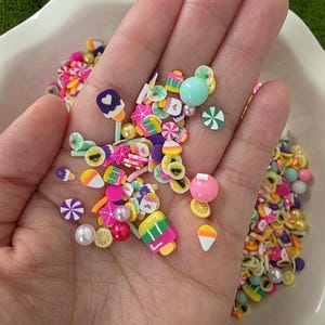 May include: A selection of colourful polymer clay sprinkles in a variety of shapes, such as ice cream cones, lollipops, and fruit slices. The sprinkles are in a white bowl, with a green moss background. The sprinkles are in shades of pink, yellow, green, and purple.
