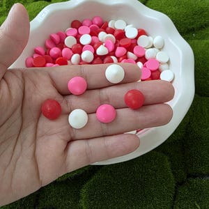 May include: A white, ruffled bowl filled with red, pink, and white candy discs. Six candies are held in a hand, showcasing the colors and shapes. The candies are a mix of red, pink, and white, with a flat, disc-like shape. The background is a green, textured surface.