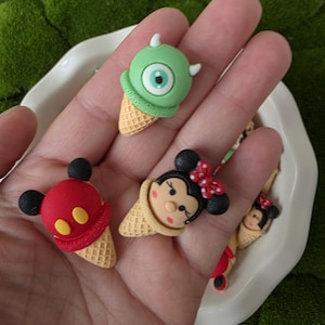 May include: A collection of colorful, cartoon-themed ice cream cone charms. The charms feature a green monster with one eye, a red Mickey Mouse, and a Minnie Mouse with a red bow. The ice cream cones are a waffle cone color.