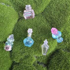 Peut inclure: Cinq bouteilles miniatures en verre de formes et de couleurs variées. Une bouteille rose carrée avec une étiquette, une bouteille bleue ronde avec une étiquette, une bouteille transparente en forme de goutte, une petite bouteille bleue avec un bouchon rose et une bouteille transparente avec une étiquette.
