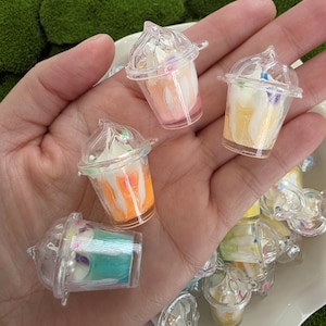 May include: Small clear plastic dessert cups filled with layers of orange, pink, yellow, and blue, topped with white cream and sprinkles. The cups have clear lids. The desserts are on a white plate.
