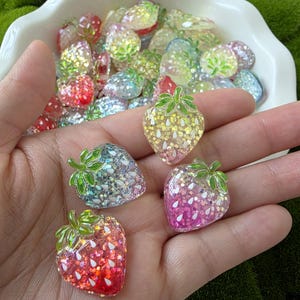 以下が含まれることがあります： キラキラ光るイチゴ型の装飾品をいくつか手に持っています。イチゴは赤、ピンク、黄色、青など様々な色で、緑の葉のディテールがあります。背景には、同様の装飾品が入った白いボウルがあります。