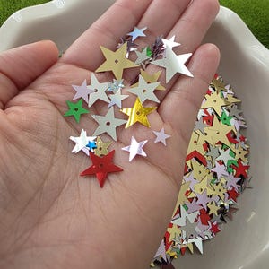 May include: A hand holding a collection of star-shaped confetti in various colors, including gold, silver, red, and green. The confetti is scattered over a white, scalloped-edge dish, with a green, moss-like background.