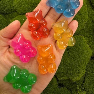May include: A collection of colorful gummy bear-shaped candies in shades of red, orange, yellow, pink, blue, and green. The candies are displayed on a bed of green moss, showcasing their translucent, glossy appearance.