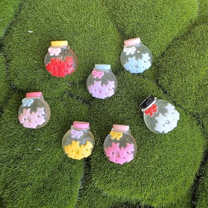 May include: Seven small, round, clear jars with colorful contents and lids. Each jar has a different color scheme, including red, pink, yellow, blue, and purple. The jars are decorated with star-shaped accents and bows.