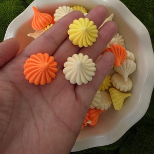 以下が含まれることがあります： 手には、オレンジ、黄色、クリーム色の小さな花や渦巻きの形をした、カラフルな樹脂製の装飾品が集められています。白い、スカラップエッジの皿に並べられています。