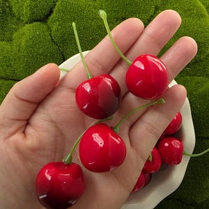 Puede incluir: Cerezas rojas brillantes con tallos verdes se muestran en un cuenco blanco y se sostienen en una mano. Las cerezas tienen un acabado brillante. El fondo es una superficie verde texturizada. Estas cerezas son un artículo decorativo.